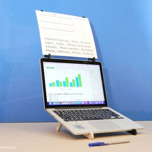 Op de afbeelding: Een zilveren laptop met een wit toetsenbord staat op een houten standaard. Een vel papier is met zwarte clips aan de bovenkant van de laptop bevestigd. Het papier bevat een lijst met notities en herinneringen. Een blauwe pen ligt op het oppervlak.