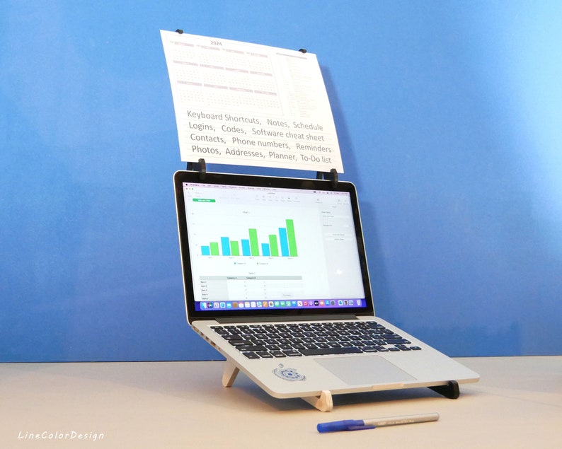 Op de afbeelding: Een zilveren laptop met een wit toetsenbord staat op een houten standaard. Een vel papier is met zwarte clips aan de bovenkant van de laptop bevestigd. Het papier bevat een lijst met notities en herinneringen. Een blauwe pen ligt op het oppervlak.
