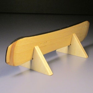 May include: A wooden snowboard model with a black top edge, displayed on a light-colored wooden stand. The snowboard has a natural wood grain finish and is supported by two triangular stands. The design includes drilled holes.
