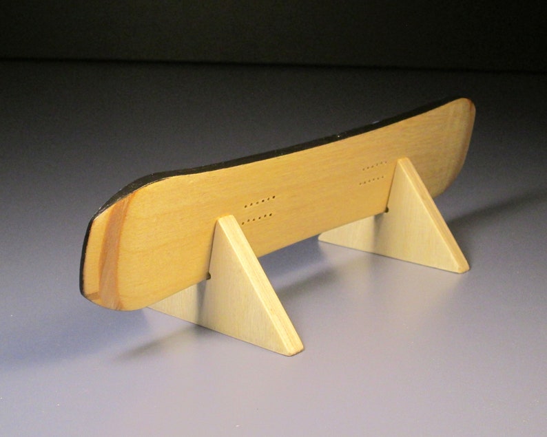 May include: A wooden snowboard model with a black top edge, displayed on a light-colored wooden stand. The snowboard has a natural wood grain finish and is supported by two triangular stands. The design includes drilled holes.