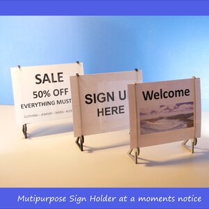 Op de afbeelding: Drie kleine, witte borden met zwarte tekst op een wit oppervlak tegen een blauwe achtergrond. De borden luiden "SALE 50% OFF", "SIGN UP HERE" en "Welcome". De borden worden vastgehouden door kleine, donkere metalen standaards. De tekst "Mutipurpose Sign Holder at a moments notice" staat onderaan.