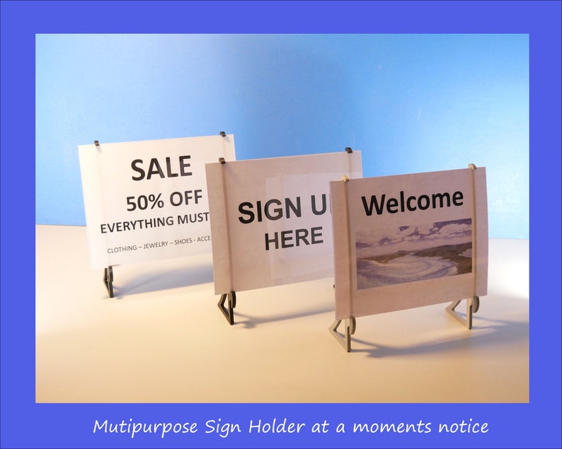 Op de afbeelding: Drie kleine, witte borden met zwarte tekst op een wit oppervlak tegen een blauwe achtergrond. De borden luiden "SALE 50% OFF", "SIGN UP HERE" en "Welcome". De borden worden vastgehouden door kleine, donkere metalen standaards. De tekst "Mutipurpose Sign Holder at a moments notice" staat onderaan.