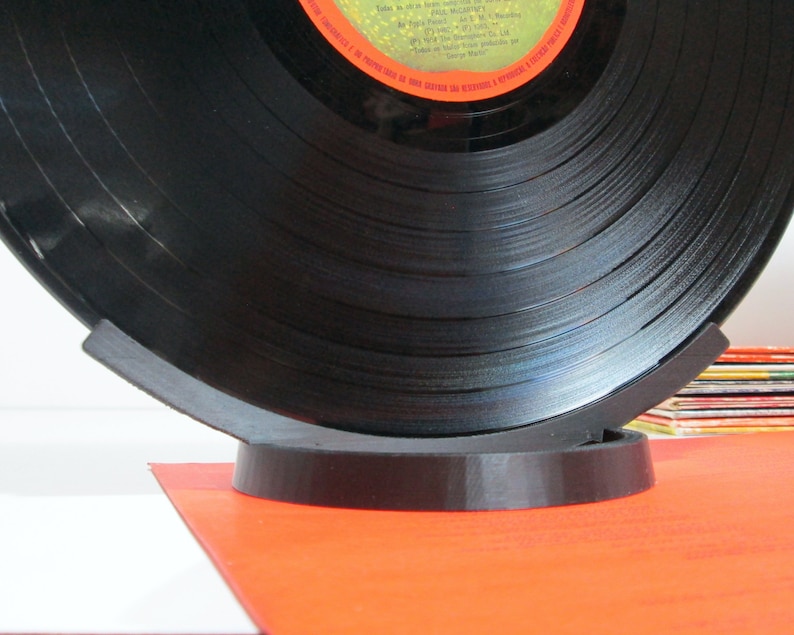 May include: A black vinyl record displayed on a dark gray stand. The record is partially visible, showing its grooves and label. The stand has a curved design, supporting the record. The background includes a stack of colorful album covers.