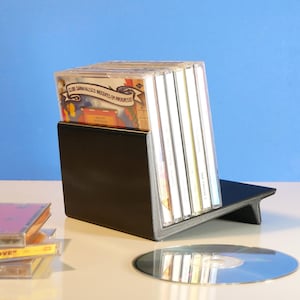 May include: A black CD holder with a slanted design, holding several CD cases. A few CDs and a stack of CD cases are on the table. The background is a light blue.