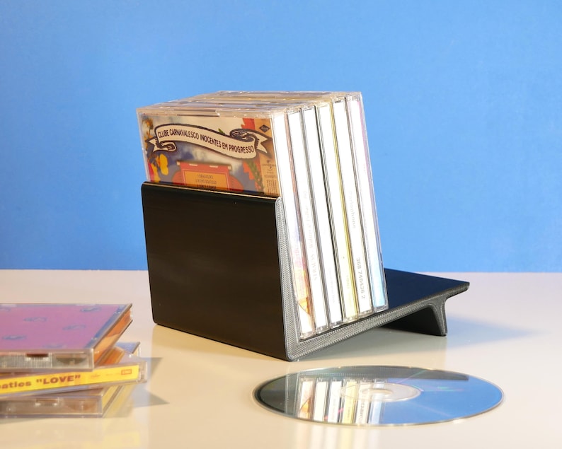 May include: A black CD holder with a slanted design, holding several CD cases. A few CDs and a stack of CD cases are on the table. The background is a light blue.