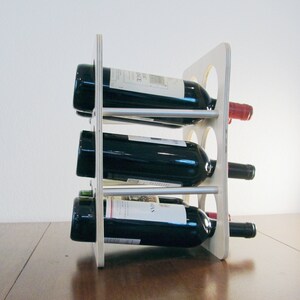 May include: A wooden wine rack holding three bottles of red wine. The rack is made of light-colored wood with circular cutouts and silver metal supports. The wine bottles are dark with white labels.