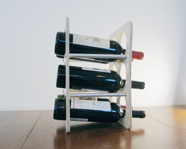 May include: A wooden wine rack holding three bottles of red wine. The rack is made of light-colored wood with circular cutouts and silver metal supports. The wine bottles are dark with white labels.