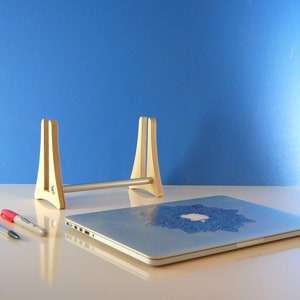 May include: A light wooden laptop stand with a silver bar, holding a silver laptop with a blue mandala design. Two pens and a marker are on the table. The background is a solid blue.