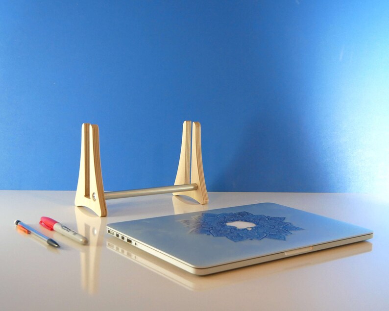 May include: A light wooden laptop stand with a silver bar, holding a silver laptop with a blue mandala design. Two pens and a marker are on the table. The background is a solid blue.
