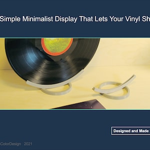 May include: A minimalist vinyl record display stand in light gray. The stand is designed to hold a vinyl record, showcasing its label. The design is simple and modern, with the text "Designed and Made in USA".