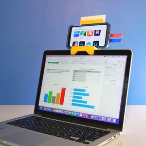 May include: A yellow smartphone holder attached to a laptop, displaying a spreadsheet. The smartphone shows app icons. The laptop screen displays a bar chart. The background is a solid blue.