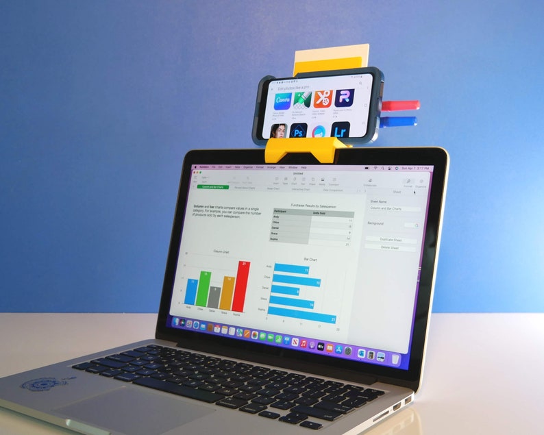 May include: A yellow smartphone holder attached to a laptop, displaying a spreadsheet. The smartphone shows app icons. The laptop screen displays a bar chart. The background is a solid blue.
