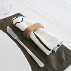 May include: A table setting featuring a white napkin with black stripes, held by a wooden ring, resting on a brown placemat. A stainless steel butter knife and a white plate are also present, suggesting a dining theme.