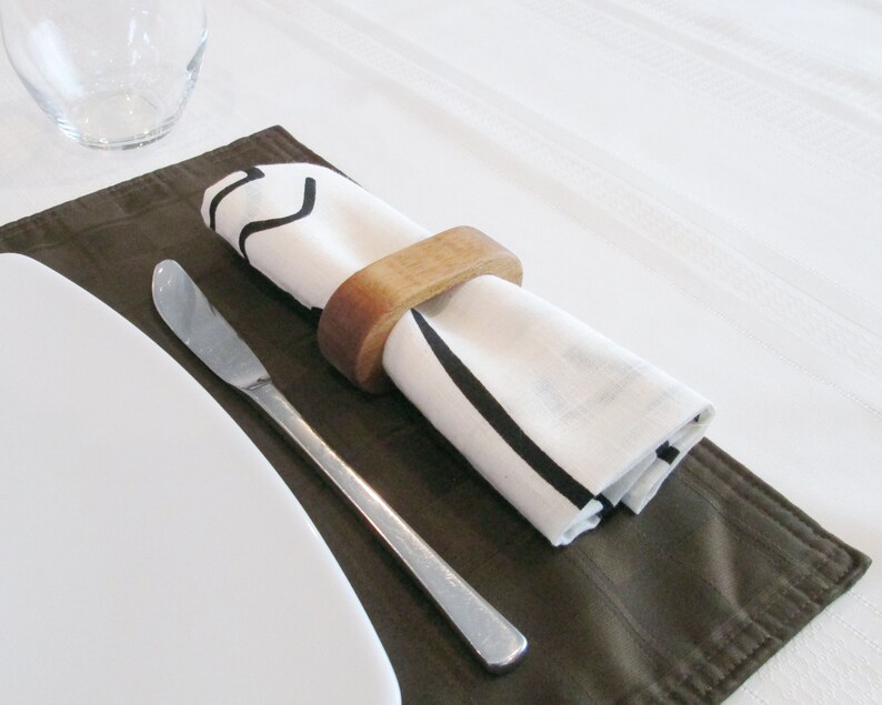 May include: A table setting featuring a white napkin with black stripes, held by a wooden ring, resting on a brown placemat. A stainless steel butter knife and a white plate are also present, suggesting a dining theme.