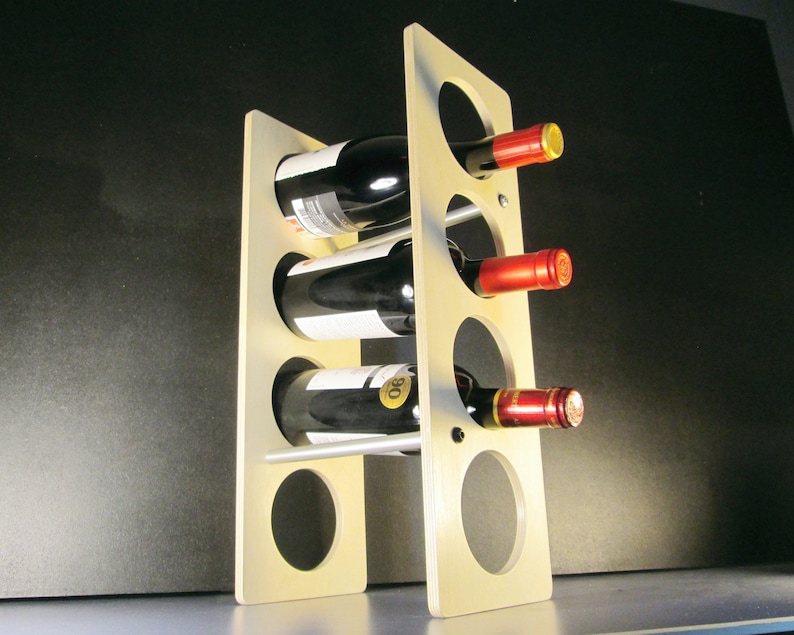 May include: A light-colored wooden wine rack holding three bottles of red wine. The rack has a modern design with circular openings to hold the bottles. The wine bottles have red and gold foil on the top.