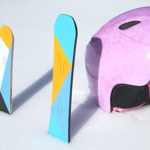 May include: Two colorful snowboards with geometric designs in yellow, blue, and white stand upright in the snow next to a pink ski helmet. The snowboards have a wooden core and black edges. The helmet has a floral pattern.
