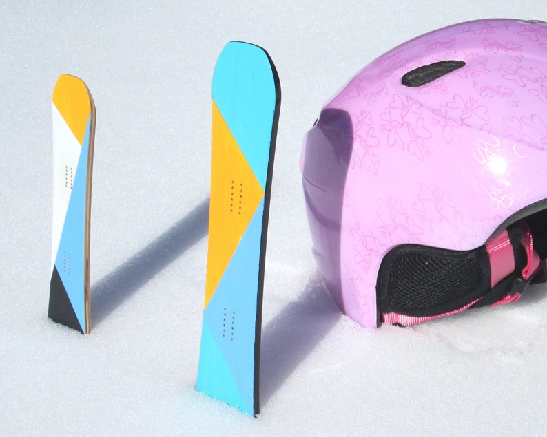 May include: Two colorful snowboards with geometric designs in yellow, blue, and white stand upright in the snow next to a pink ski helmet. The snowboards have a wooden core and black edges. The helmet has a floral pattern.