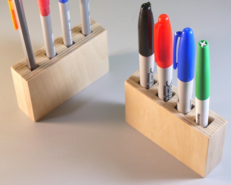 May include: Two wooden pen holders, each with four slots, are displayed. One holds a pencil and three pens, while the other holds four Sharpie markers in black, red, blue, and green. The holders are made of light-colored wood.