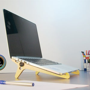 May include: A laptop computer rests on a light-colored wooden stand. The stand is angled to elevate the laptop. A blue pen and a pen holder with colorful pens are visible. The image includes a logo that says "Line Color Design".