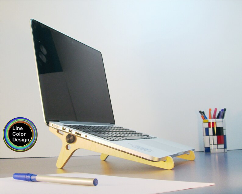 May include: A laptop computer rests on a light-colored wooden stand. The stand is angled to elevate the laptop. A blue pen and a pen holder with colorful pens are visible. The image includes a logo that says "Line Color Design".