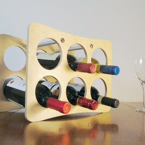 May include: A light-colored wooden wine rack with six circular openings, each holding a wine bottle. The rack has a curved design and is on a wooden surface. A wine glass sits nearby.