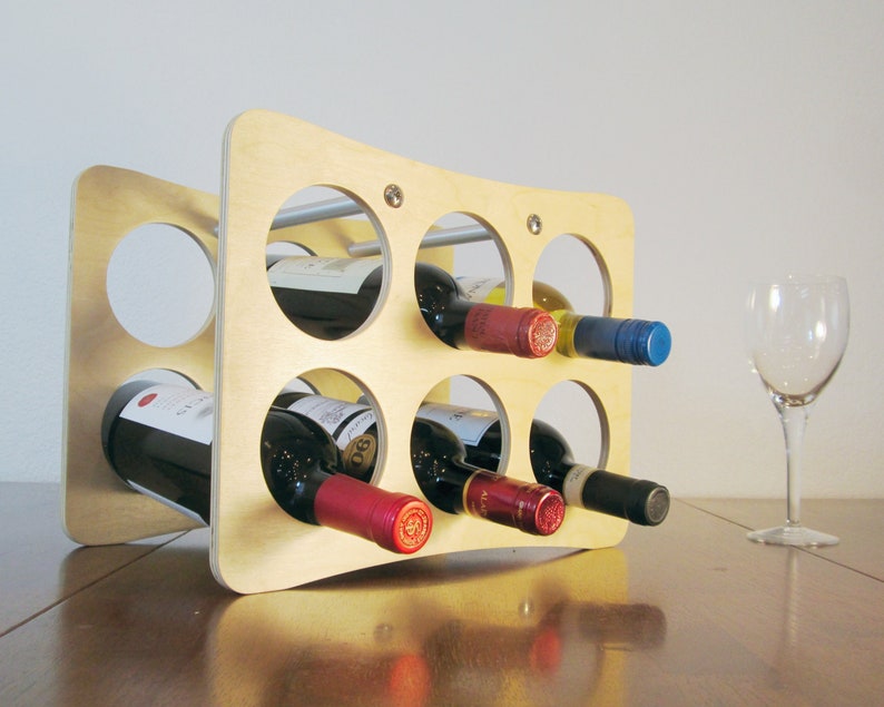 May include: A light-colored wooden wine rack with six circular openings, each holding a wine bottle. The rack has a curved design and is on a wooden surface. A wine glass sits nearby.