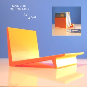 May include: An orange and yellow CD holder with a modern design. The holder is made in Colorado by Alan. The holder is designed to hold multiple CDs. A CD is placed on the surface next to the holder.