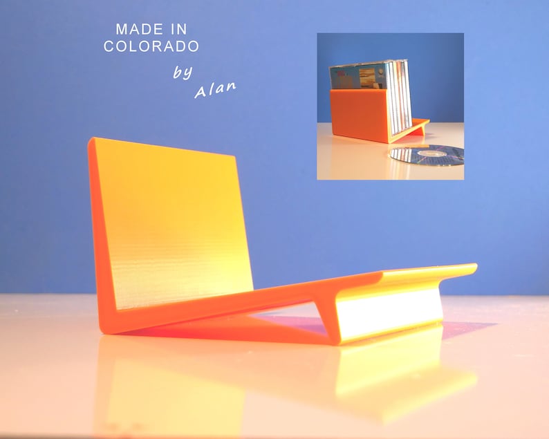 May include: An orange and yellow CD holder with a modern design. The holder is made in Colorado by Alan. The holder is designed to hold multiple CDs. A CD is placed on the surface next to the holder.