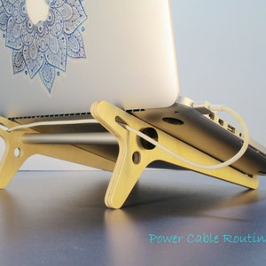 May include: A light-colored wooden laptop stand with a silver laptop resting on it. The stand has a unique design with cut-out shapes and a horizontal bar. A white power cable is connected to the laptop. The text "Power Cable Routing" is visible.