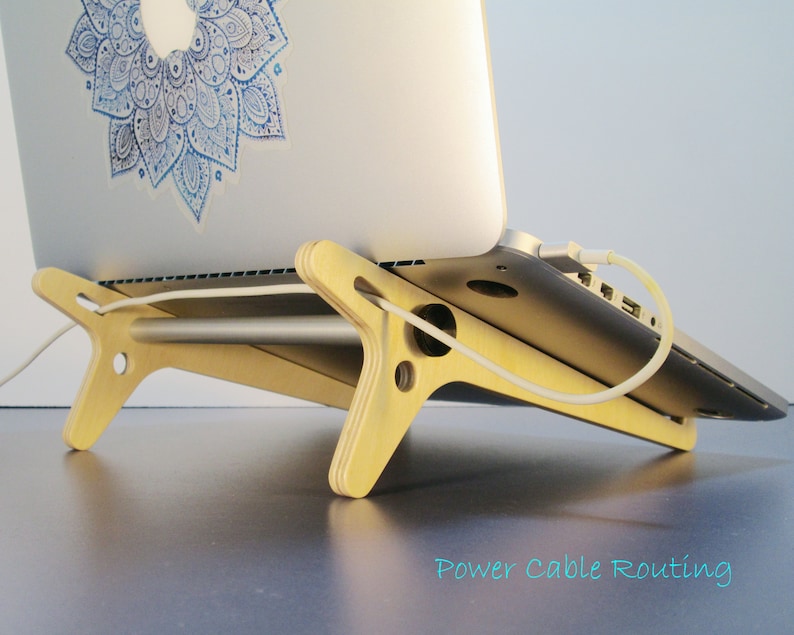 May include: A light-colored wooden laptop stand with a silver laptop resting on it. The stand has a unique design with cut-out shapes and a horizontal bar. A white power cable is connected to the laptop. The text "Power Cable Routing" is visible.