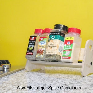 May include: A light-colored wooden spice rack holding several spice jars, including black pepper, Italian seasoning, oregano, and garlic powder. The rack is on a countertop with a yellow background. Text on the image reads "Also Fits Larger Spice Containers."