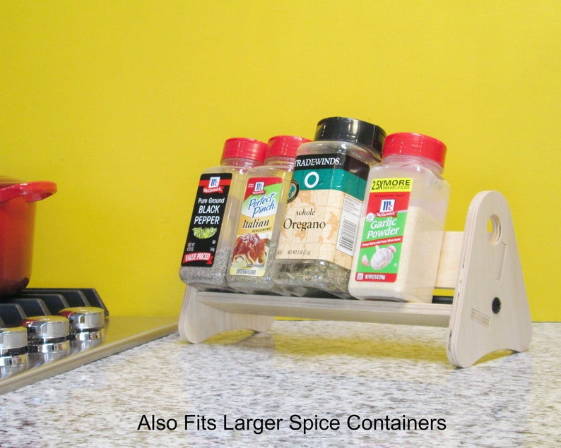 May include: A light-colored wooden spice rack holding several spice jars, including black pepper, Italian seasoning, oregano, and garlic powder. The rack is on a countertop with a yellow background. Text on the image reads "Also Fits Larger Spice Containers."