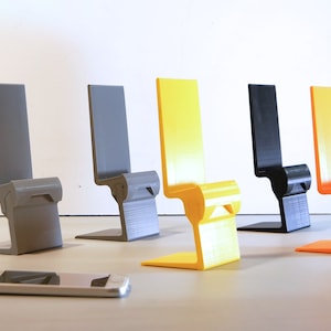 K&ouml;nnte beinhalten: Eine Sammlung von f&uuml;nf farbenfrohen Telefonst&auml;ndern in Grau, Gelb, Schwarz und Orange. Jeder St&auml;nder hat ein einzigartiges stuhl&auml;hnliches Design mit einem Sockel und einer R&uuml;ckenlehne. Ein Smartphone liegt vor den St&auml;ndern.