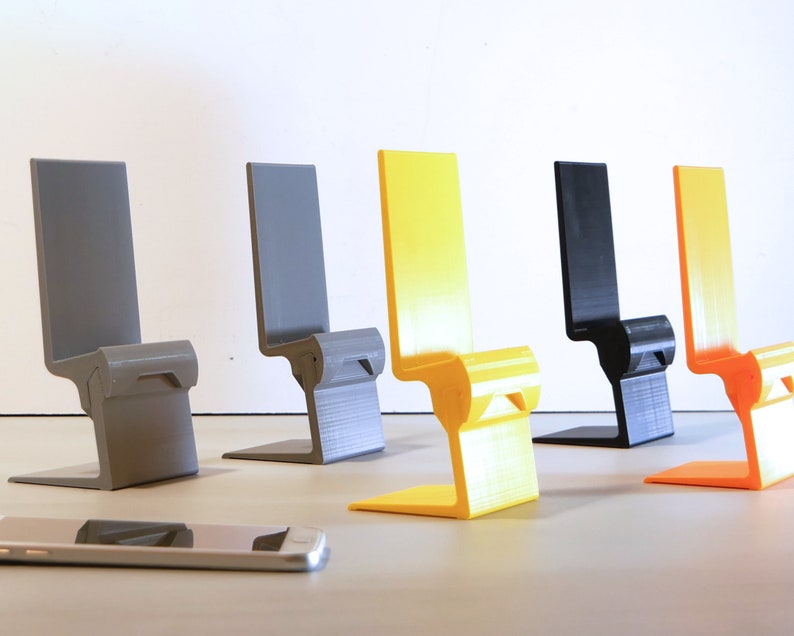 K&ouml;nnte beinhalten: Eine Sammlung von f&uuml;nf farbenfrohen Telefonst&auml;ndern in Grau, Gelb, Schwarz und Orange. Jeder St&auml;nder hat ein einzigartiges stuhl&auml;hnliches Design mit einem Sockel und einer R&uuml;ckenlehne. Ein Smartphone liegt vor den St&auml;ndern.