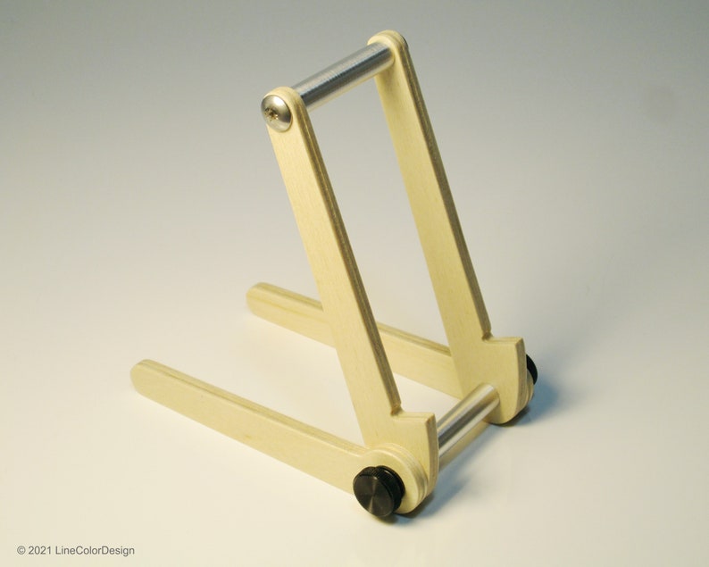 May include: A light-colored wooden stand with a metal bar and black hardware. The stand is designed to hold a tablet or book. The design is simple and functional, with a minimalist aesthetic. The stand is made of wood and metal.