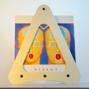 May include: A light-coloured wooden record album display stand in a triangular shape, with black fixings. The stand holds a record album with a colourful abstract design. The word "DIRECT" is visible on the album cover.