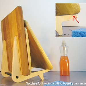 May include: A wooden cutting board stand with a light wood frame and a dark wood cutting board. The stand has notches for holding the cutting board at an angle to dry. The stand is made of light-colored wood and has a black screw.