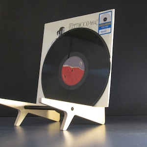 May include: A black vinyl record in a white album cover, displayed on a light wood stand. The album cover features the band name "Fleetwood Mac" and a photo. The record has a red and purple label. An "Exclusive Clear Vinyl" sticker is visible.