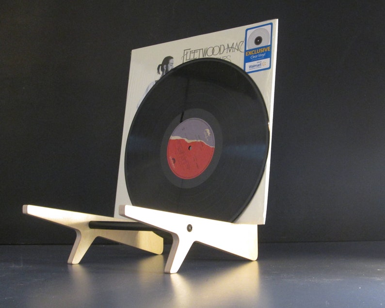 May include: A black vinyl record in a white album cover, displayed on a light wood stand. The album cover features the band name "Fleetwood Mac" and a photo. The record has a red and purple label. An "Exclusive Clear Vinyl" sticker is visible.