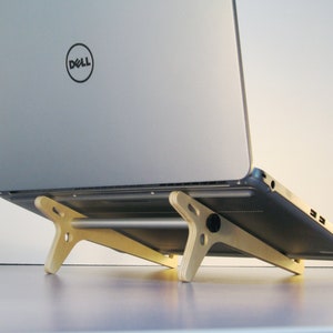 May include: A silver laptop with a Dell logo is elevated on a light-colored wooden laptop stand. The stand has a minimalist design with angled legs and a horizontal support bar, providing an ergonomic setup.