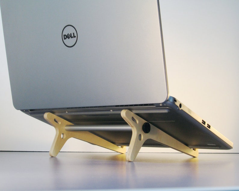 May include: A silver laptop with a Dell logo is elevated on a light-colored wooden laptop stand. The stand has a minimalist design with angled legs and a horizontal support bar, providing an ergonomic setup.