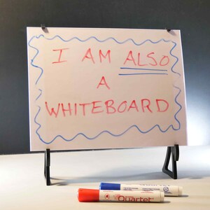 Op de afbeelding: Een wit whiteboard met een blauwe golvende rand en de tekst "I AM ALSO A WHITEBOARD" in rode marker. Het whiteboard staat op een zwarte standaard, met rode, blauwe en witte stiften ervoor.