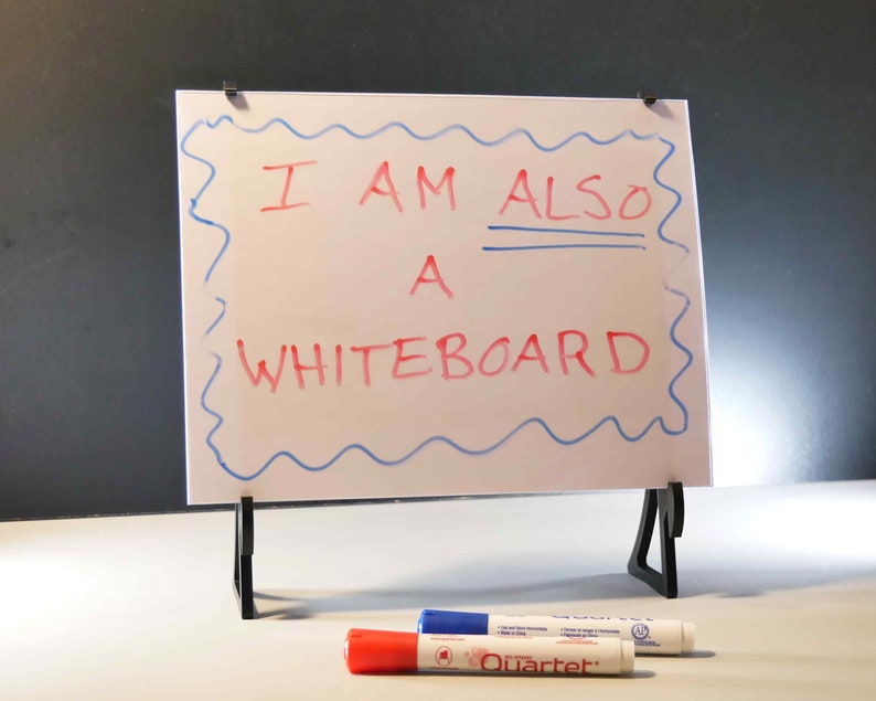 Op de afbeelding: Een wit whiteboard met een blauwe golvende rand en de tekst "I AM ALSO A WHITEBOARD" in rode marker. Het whiteboard staat op een zwarte standaard, met rode, blauwe en witte stiften ervoor.