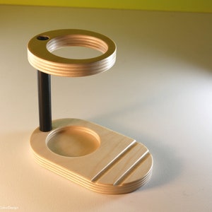 May include: A wooden desk organizer with a light wood finish. It features a circular top with a small hole, a black support rod, and an oval base with a recessed area and three angled grooves. The design is modern and minimalist.