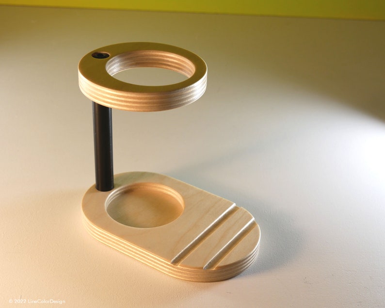 May include: A wooden desk organizer with a light wood finish. It features a circular top with a small hole, a black support rod, and an oval base with a recessed area and three angled grooves. The design is modern and minimalist.