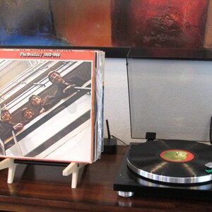 May include: A stack of vinyl records in a wooden display stand, with the album cover of The Beatles' 1962-1966 album visible. A record player with a black vinyl record is in the foreground. The album cover features a photo of the band.