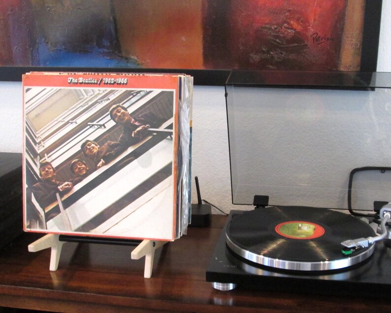 May include: A stack of vinyl records in a wooden display stand, with the album cover of The Beatles' 1962-1966 album visible. A record player with a black vinyl record is in the foreground. The album cover features a photo of the band.