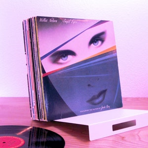 May include: A collection of vinyl records, including Willie Nelson's "Angel Eyes" album, is displayed on a white, rectangular stand. The album cover shows a woman's face with blue eyes. A single record is in the foreground.