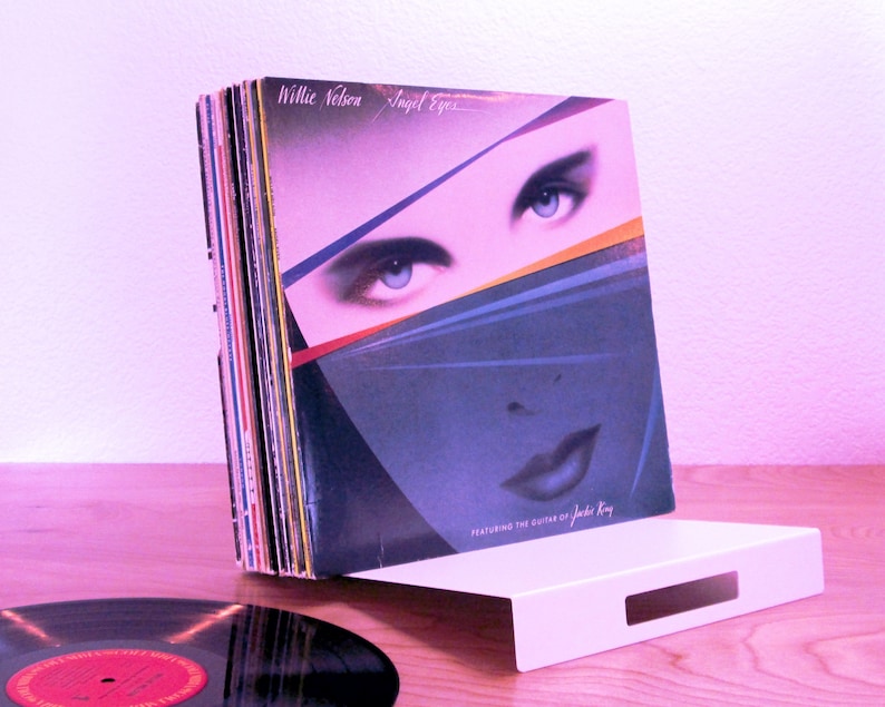 May include: A stack of vinyl records, including Willie Nelson's "Angel Eyes" album, rests on a white, rectangular stand. The album cover features a woman's face with blue eyes. A single record is visible in the foreground.