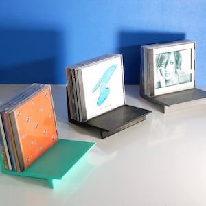 May include: Three CD holders in teal, black, and silver, each holding a stack of CDs. The teal holder is on the left, the black in the middle, and the silver on the right. The CDs have various album art, including a blue design and a portrait.
